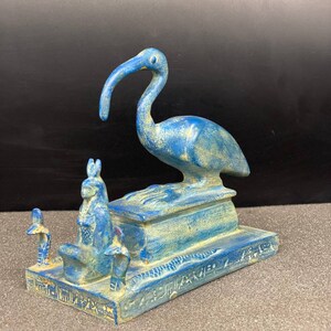 Puede incluir: Figurilla pintada en azul y dorado del dios egipcio Thoth, representado como un pájaro Ibis de pie sobre un pedestal con jeroglíficos.
