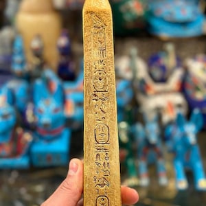 Puede incluir: Un obelisco dorado y alargado con jeroglíficos tallados. El obelisco, sostenido en una mano, tiene una parte superior puntiaguda. Otras figuras de temática egipcia son visibles en el fondo borroso.