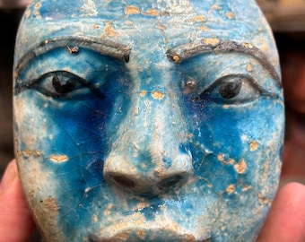 May include: A blue ceramic face mask with black eyes, eyebrows, and lips. The mask has a cracked glaze effect and is held in a person's hand. The mask is a light blue color with darker blue accents.