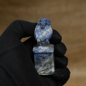 May include: A lapis lazuli ram figurine with a detailed carving. The ram is perched atop a rectangular base with hieroglyphic carvings. The figurine is held in a black gloved hand against a brown background.