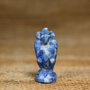 May include: A small, blue and white stone sculpture of a ram. The ram sculpture is carved from a stone with a marbled appearance. The sculpture is standing on a small base. The background is a neutral brown.