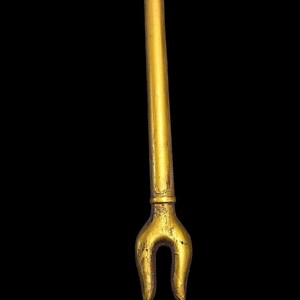 Puede incluir: Un objeto de metal de color dorado con un eje largo y delgado y un extremo bifurcado. El objeto recuerda a un bast&oacute;n o cetro ceremonial tradicional.