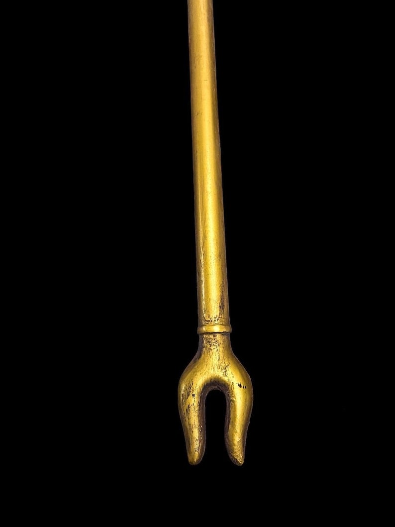 Puede incluir: Un objeto de metal de color dorado con un eje largo y delgado y un extremo bifurcado. El objeto recuerda a un bast&oacute;n o cetro ceremonial tradicional.