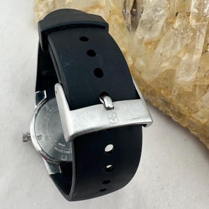 May include: Close-up of a black wristwatch with a silver buckle. The watch has a black rubber strap and a silver case. The watch face is visible, with a white background and black markings. The watch is water resistant.