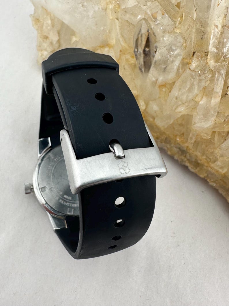May include: Close-up of a black wristwatch with a silver buckle. The watch has a black rubber strap and a silver case. The watch face is visible, with a white background and black markings. The watch is water resistant.