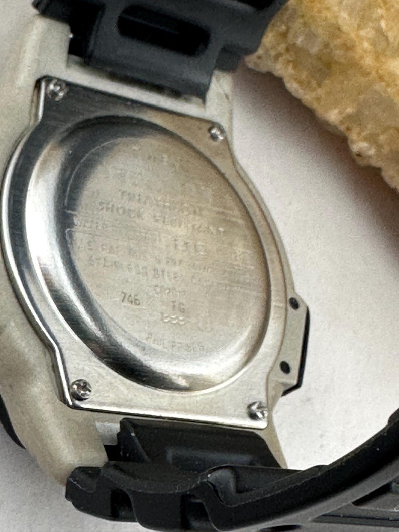 May include: Close-up of the back of a digital watch. The watch has a silver-colored metal back with engraved text and a black and white plastic band. The text includes the words "SHOCK RESISTANT" and "STAINLESS STEEL".