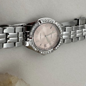 May include: A silver Guess wristwatch with a pale pink face and a bezel set with small, clear crystals. The watch features a date display and a silver link bracelet. The watch is shown against a light background.