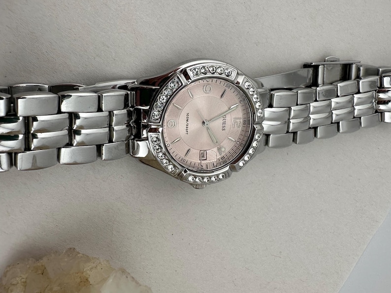 May include: A silver Guess wristwatch with a pale pink face and a bezel set with small, clear crystals. The watch features a date display and a silver link bracelet. The watch is shown against a light background.