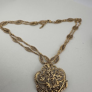 May include: Gold-toned filigree necklace featuring a heart-shaped pendant with floral and scroll details. The pendant has dangling accents. The necklace chain has an oval link design. The jewelry is displayed on a white background.