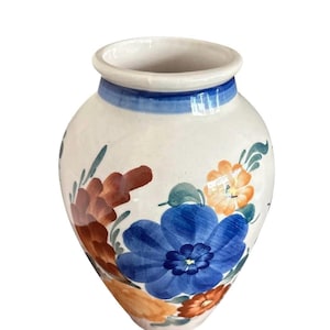 Könnte beinhalten: Eine weiße Keramikvase mit rundem Körper und blauem Rand. Die Vase ist mit handgemalten Blumen in Blau-, Orange- und Brauntönen verziert. Das florale Design verleiht diesem dekorativen Stück einen Hauch von rustikalem Charme.