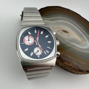 Puede incluir: Un reloj de pulsera plateado con esfera cuadrada y esfera azul oscuro. El reloj tiene subesferas blancas y naranjas, agujas rojas y visualización de fecha. La correa del reloj es de eslabones metálicos plateados.