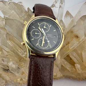 May include: A gold-coloured Seiko World Timer watch with a dark grey face and three sub-dials. The watch has a brown leather strap. The watch face displays the date and various time zones.