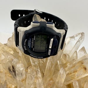 May include: A black and white Timex Ironman digital watch with a black band. The watch face displays the time and other data. The watch is water resistant and shock resistant. The watch is resting on a cluster of clear quartz crystals.