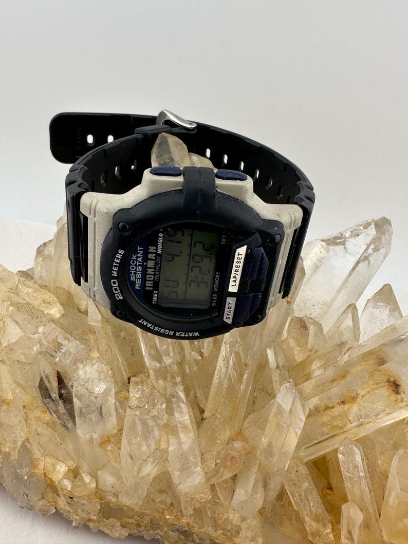 May include: A black and white Timex Ironman digital watch with a black band. The watch face displays the time and other data. The watch is water resistant and shock resistant. The watch is resting on a cluster of clear quartz crystals.