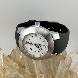 May include: A silver-toned wristwatch with a white face, black numbers, and a black rubber band. The watch face displays the text "SWISS ARMY" and "Green Guard". The watch is resting on a bed of clear quartz crystals.