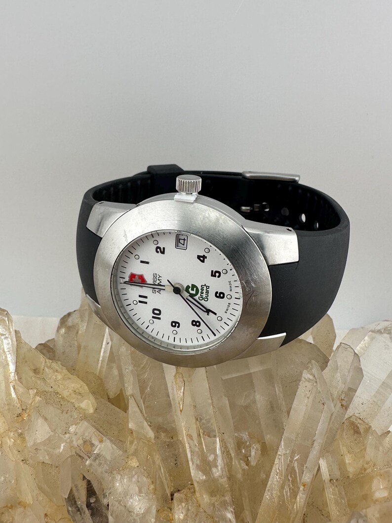 May include: A silver-toned wristwatch with a white face, black numbers, and a black rubber band. The watch face displays the text "SWISS ARMY" and "Green Guard". The watch is resting on a bed of clear quartz crystals.