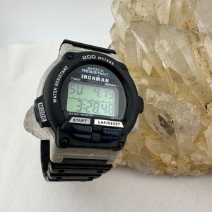 May include: Black and gray Timex Ironman digital watch. The watch displays the time and date on a digital screen. Features include water resistance, shock resistance, and a lap/reset function. The watch is set against a light background.