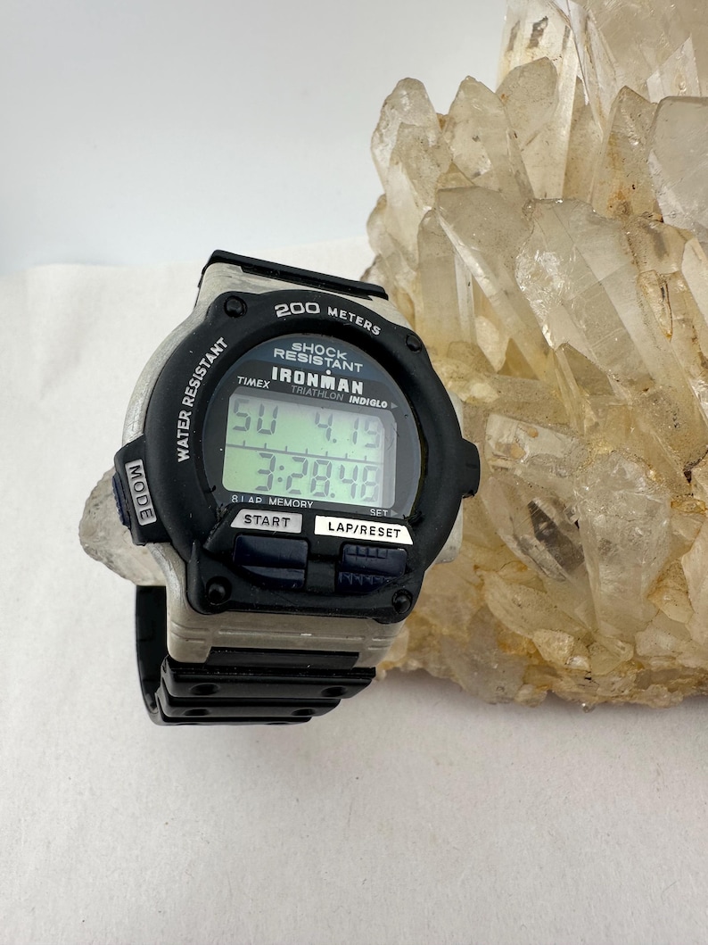 May include: Black and gray Timex Ironman digital watch. The watch displays the time and date on a digital screen. Features include water resistance, shock resistance, and a lap/reset function. The watch is set against a light background.