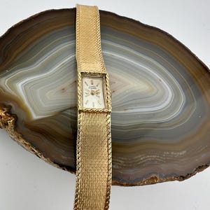 Puede incluir: Un reloj vintage dorado con una esfera rectangular y una correa tejida. La esfera del reloj es blanca con manecillas y marcadores dorados. La correa es de malla ancha y texturizada. El reloj se muestra sobre una superficie de piedra marrón y gris.