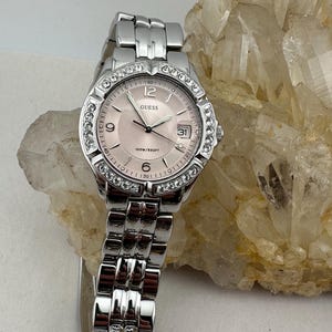 May include: A silver Guess watch with a pink face and a date display. The watch has a bezel adorned with clear crystals and a stainless steel band. The watch face displays the number 12, 9, 6, and 3. The watch is water resistant up to 100 metres.