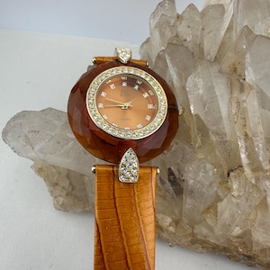 Pode incluir: Um relógio de quartzo vintage com mostrador castanho facetado e moldura dourada incrustada com cristais transparentes. O relógio tem uma pulseira de couro castanho com padrão texturizado e fecho cravejado de cristais. O mostrador tem aproximadamente 3,8 cm de diâmetro.