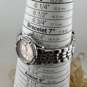 May include: A silver Guess watch with a pink face and a crystal-studded bezel. The watch is on a white measuring cone, with bracelet sizes in inches and centimetres.
