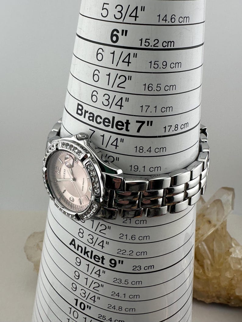 May include: A silver Guess watch with a pink face and a crystal-studded bezel. The watch is on a white measuring cone, with bracelet sizes in inches and centimetres.