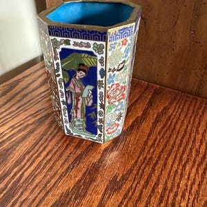 May include: An ornate, hexagonal vase with a blue interior and gold trim. The exterior features detailed enamel work with floral and figure designs in shades of blue, red, green, and white. The vase is set on a wooden surface.