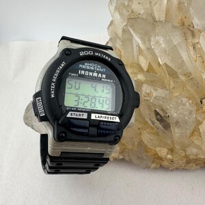 May include: A black and gray Timex Ironman digital watch with a black strap. The watch face displays the time and date. The watch is water resistant and shock resistant. The watch has the text "200 METERS" and "IRONMAN" on the face.