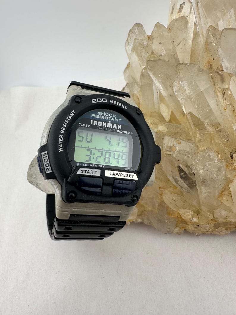 May include: A black and gray Timex Ironman digital watch with a black strap. The watch face displays the time and date. The watch is water resistant and shock resistant. The watch has the text "200 METERS" and "IRONMAN" on the face.