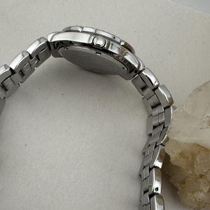 May include: A silver wristwatch with a linked metal strap. The watch face is round, and the strap curves, showing the side profile. The watch is on a light-coloured surface.