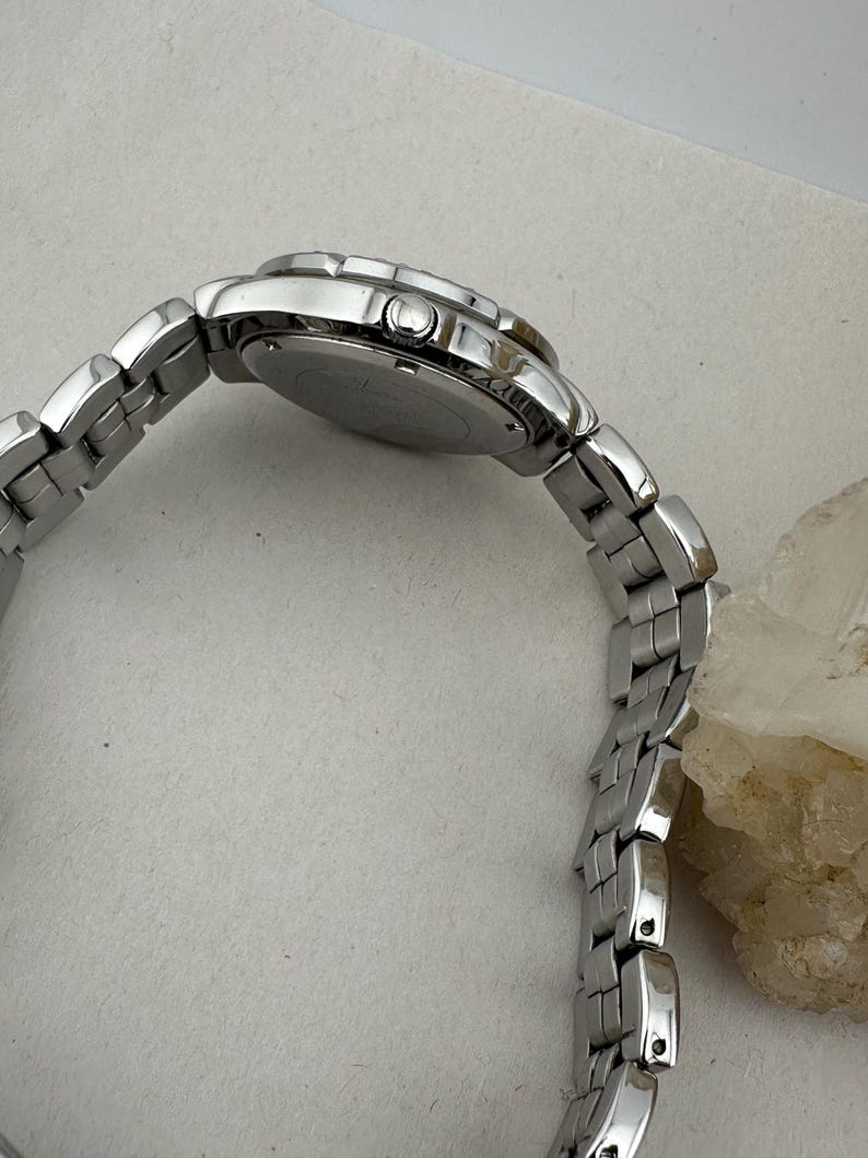 May include: A silver wristwatch with a linked metal strap. The watch face is round, and the strap curves, showing the side profile. The watch is on a light-coloured surface.