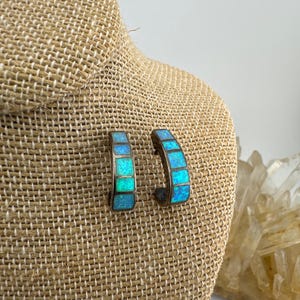 May include: A pair of hoop earrings featuring a curved metal frame inlaid with small, square-cut iridescent blue and green stones. The earrings have a segmented design with a polished metallic finish, showcasing a vibrant play of color.