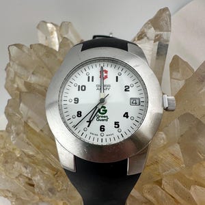 May include: A silver Swiss Army watch with a white face, black band, and the text "Green Guard." The watch has a date display at the 3 o'clock position. The watch is set against a background of clear quartz crystals.