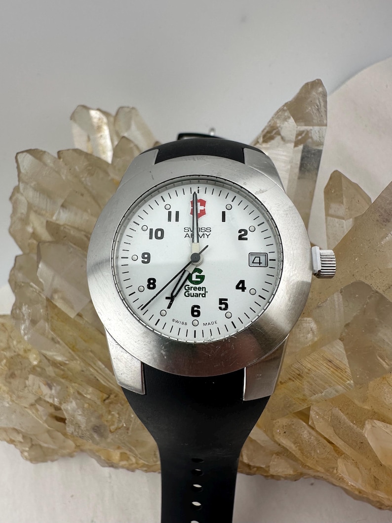 May include: A silver Swiss Army watch with a white face, black band, and the text "Green Guard." The watch has a date display at the 3 o'clock position. The watch is set against a background of clear quartz crystals.