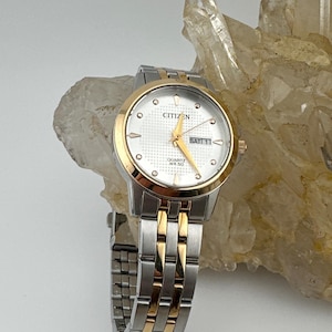 May include: A two-tone Citizen quartz watch with a silver and gold-tone stainless steel link bracelet. The round white dial features a textured center, gold-tone hands, hour markers, and a day-date window at the three o'clock position.