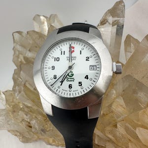 May include: A silver-toned Swiss Army watch with a white face, black hands, and a black rubber strap. The watch face features the text "Swiss Army" and "Green Guard". The watch has a date display at the 4 o'clock position.