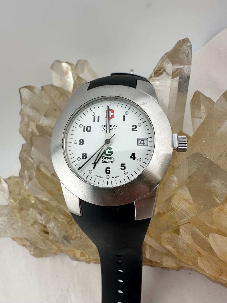 May include: A silver-toned Swiss Army watch with a white face, black hands, and a black rubber strap. The watch face features the text "Swiss Army" and "Green Guard". The watch has a date display at the 4 o'clock position.