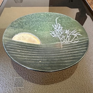Puede incluir: Un plato decorativo redondo de cerámica verde con un diseño de paisaje. El plato presenta un sol amarillo, un diseño blanco en forma de árbol y líneas blancas horizontales. El plato está sobre una mesa de cristal.