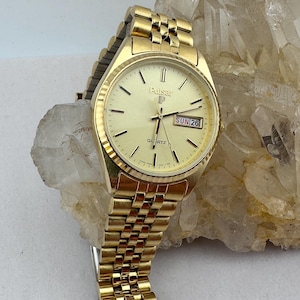 Può includere: Orologio Pulsar Quartz dorato con cinturino dorato. Il quadrante dell'orologio è di colore oro chiaro con indici e lancette neri. La data è visualizzata sul quadrante. L'orologio è su una superficie chiara.