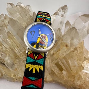 Puede incluir: Un reloj de pulsera plateado con una esfera azul que presenta una ilustración nativa americana. La correa del reloj es negra con un patrón geométrico colorido en rojo, amarillo y verde. El reloj está sobre un fondo de cristales de cuarzo transparente.
