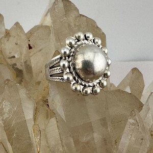 May include: A silver ring with a large, round central feature encircled by small, beaded embellishments. The ring's band has etched patterns. The ring is presented on a surface of clear quartz crystals, contrasting the shiny metal with the textured background.