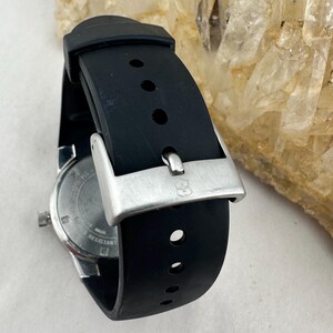 May include: A black rubber watch band with a silver buckle. The watch face is visible, showing a silver dial and the word "resistant". The band has several holes for adjustment. The watch is a classic style.