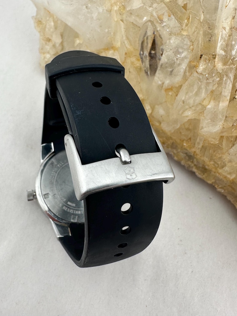 May include: A black rubber watch band with a silver buckle. The watch face is visible, showing a silver dial and the word "resistant". The band has several holes for adjustment. The watch is a classic style.