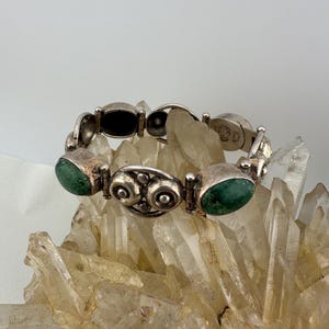 Op de afbeelding: Zilverkleurige armband met ovale schakels, met groene en zwarte stenen accenten. De armband heeft een decoratief ontwerp op sommige schakels en wordt gepresenteerd op heldere kristallen. Een vintage stuk.