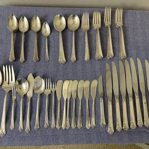 May include: A collection of vintage silver-plated flatware, including spoons, forks, and knives, arranged on a blue tablecloth. The silverware features ornate floral detailing on the handles. The set includes various sizes of spoons, forks, and knives.