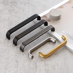 May include: A collection of cabinet handles in various metallic finishes: black, dark gray, silver, and gold. Each handle features a curved design with a ridged texture, showcasing a modern aesthetic. The handles are arranged on a light gray surface.