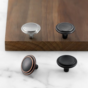 May include: Four cabinet knobs in different finishes: nickel, black, oil rubbed bronze, and black with a copper accent. The knobs are round with a raised edge.