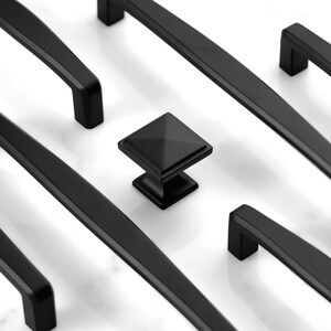 May include: A collection of matte black cabinet hardware including several long, straight drawer pulls and a single square-shaped cabinet knob with a pyramid-style top, all arranged in a geometric pattern on a white surface.