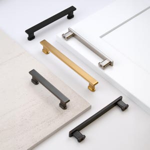 May include: An assortment of cabinet pulls in various finishes, including matte black, brushed nickel, gold, and bronze. The pulls are rectangular with rounded ends, displayed against a white cabinet door and a textured surface.